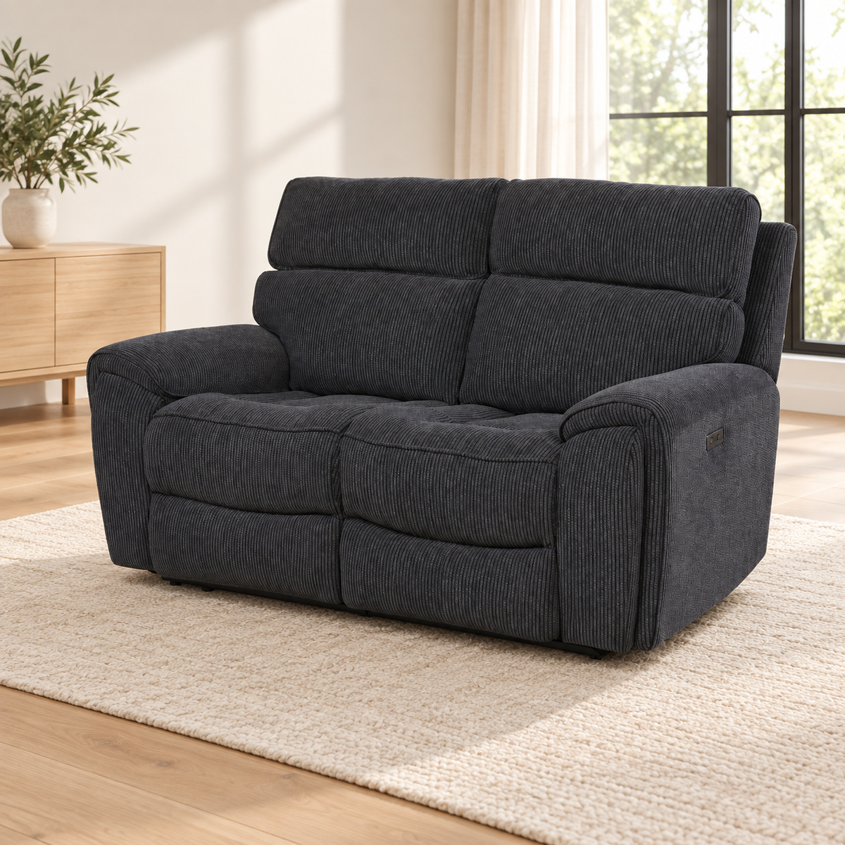 A dark charcoal gray corduroy reclining loveseat sofa placed on a light beige woven rug in a bright, modern living room with wood floors, a sideboard, and a window.