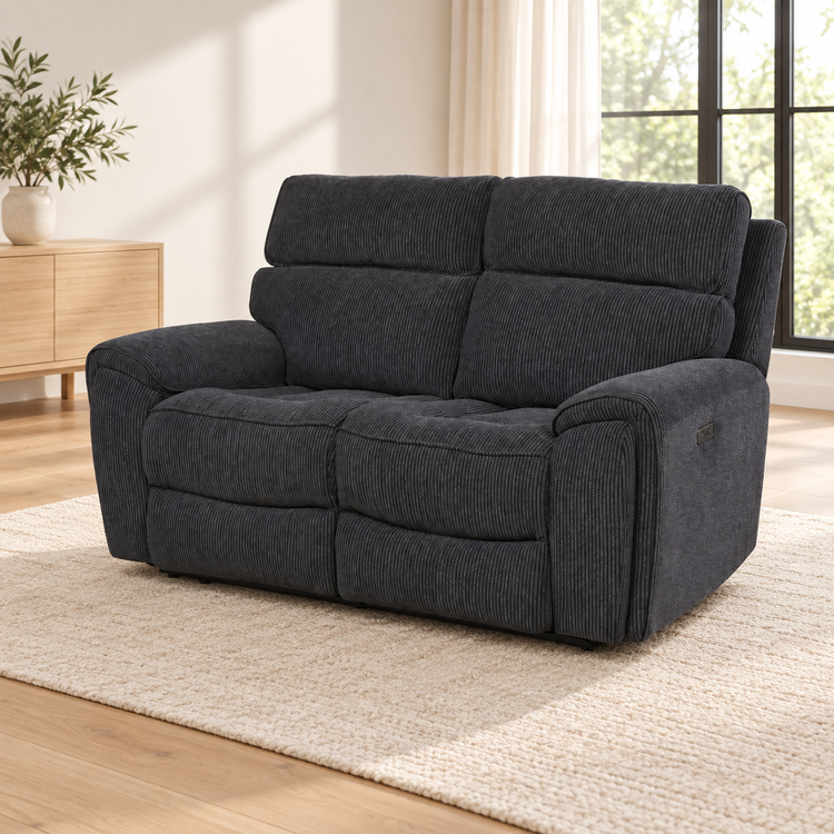 A dark charcoal gray corduroy reclining loveseat sofa placed on a light beige woven rug in a bright, modern living room with wood floors, a sideboard, and a window.