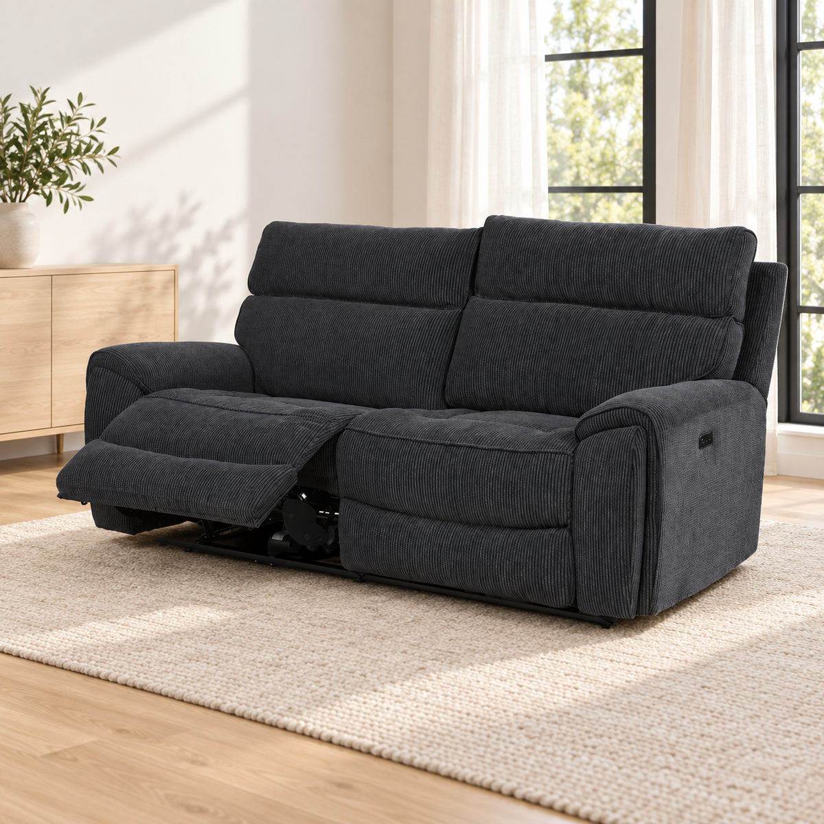 A dark charcoal gray reclining loveseat sofa shown in a bright, modern living room setting with a light wood floor, beige rug, and window in the background.